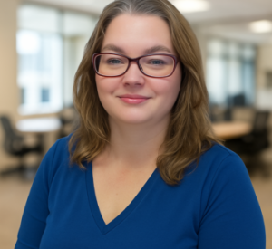 Marsha, founder of Always Typing, wearing a blue V-neck sweater and glasses, seated in front of a softly blurred office background.